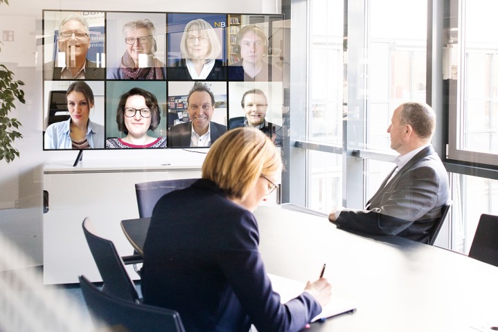 Eine Frau und ein Mann am Schreibtisch vor einem Monitor in einer Videokonferenz. Im Monitor werden Gesichter von anderen Menschen gezeigt.